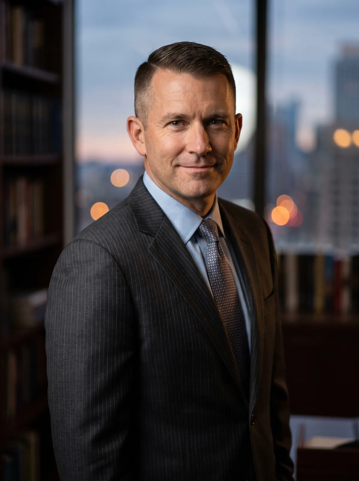 Daniel R. Kovar in a dark pinstripe suit, standing in his office in front of a wall of law books, city lights behind him at dusk